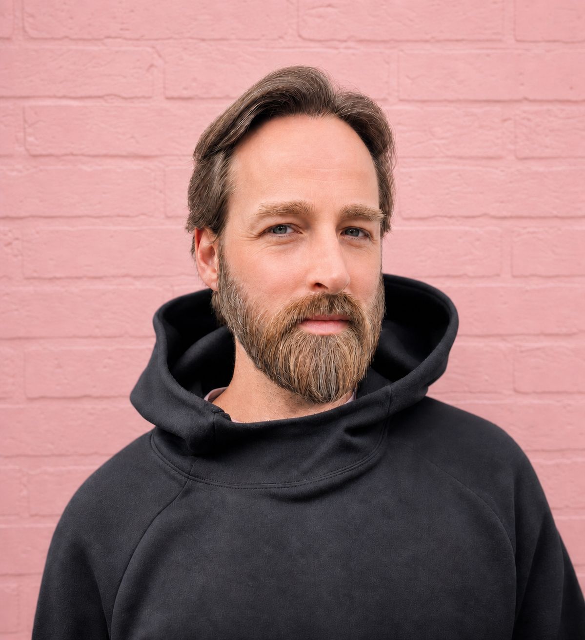 Picture of John Archbold wearing a black hoodie, outside, with a pink brickwork background